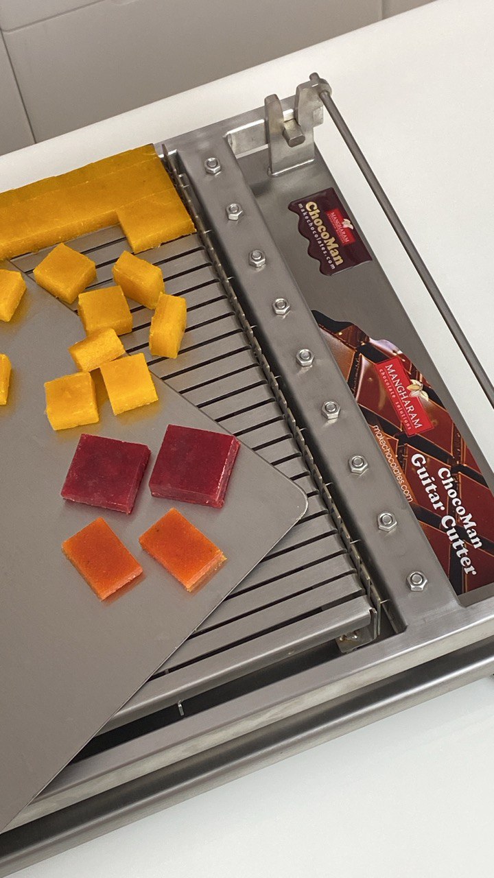 ChocoMan Guitar cutter with sliced cubes of aam papad/aam shotto or mango fruit leather roll-ups on a white surface, manufactured in India